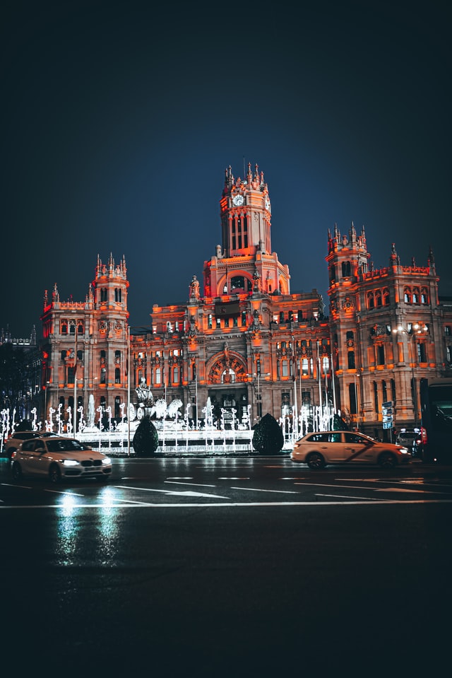 Palacio de Cibeles illuminated against a dark blue sky, with a fountain and cars in the foreground